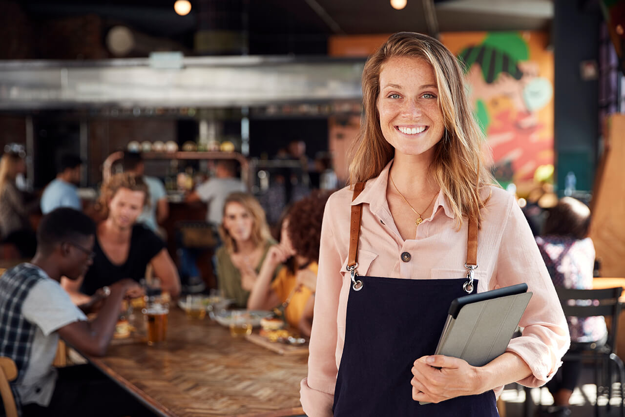 Die Inhaberin eines Restaurants steht freudestrahlend in ihrem gut besuchten Restaurant und schaut uns an.